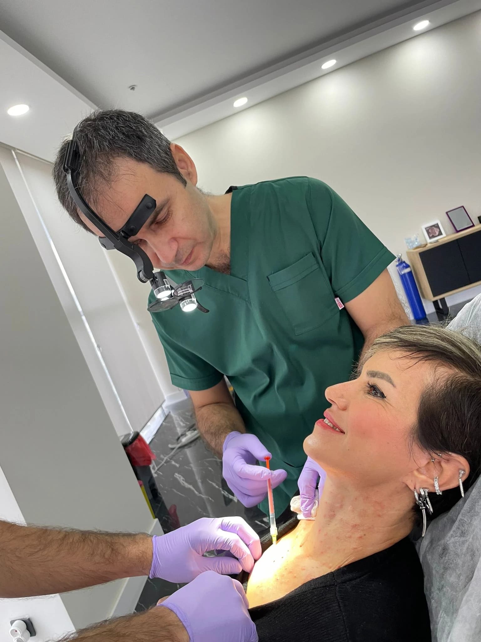 A doctor wearing surgical loupes injecting a patient's neck during a cosmetic procedure.