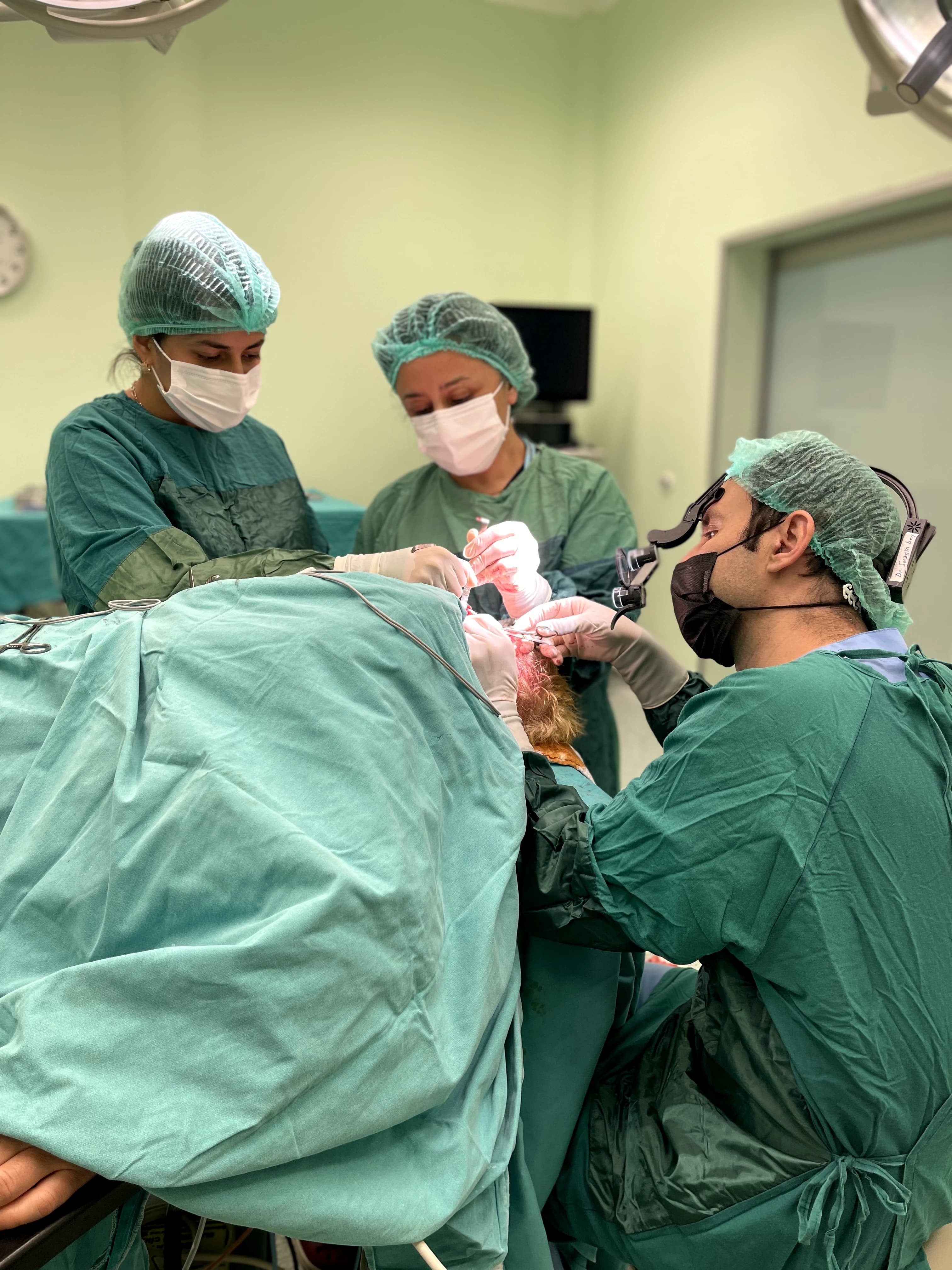 Three surgeons in green surgical gowns and caps perform a surgery on a patient under bright surgical lights in an operating room.