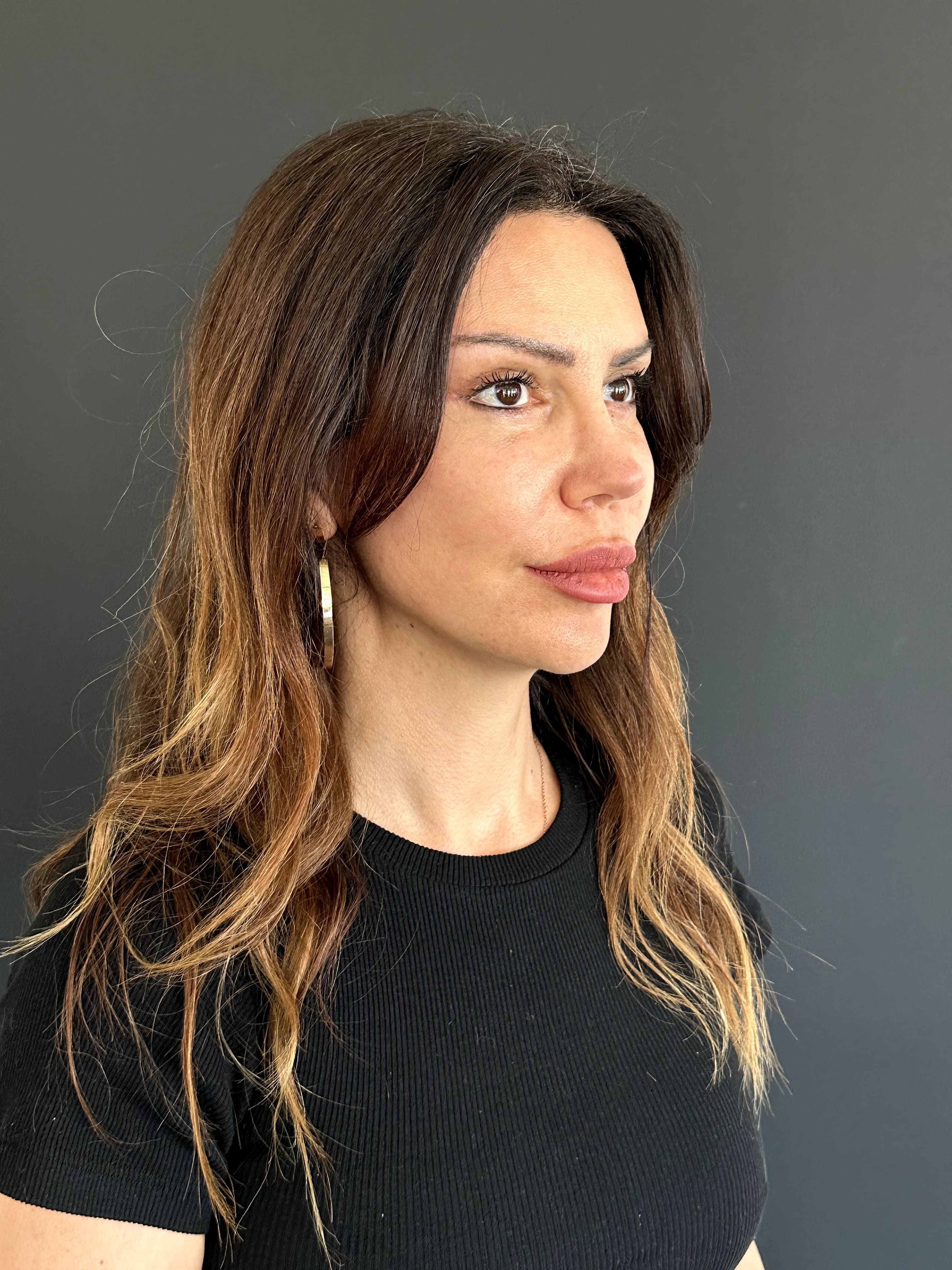Side profile of a woman with long, highlighted hair, wearing a black shirt and gold hoop earrings, against a gray background.