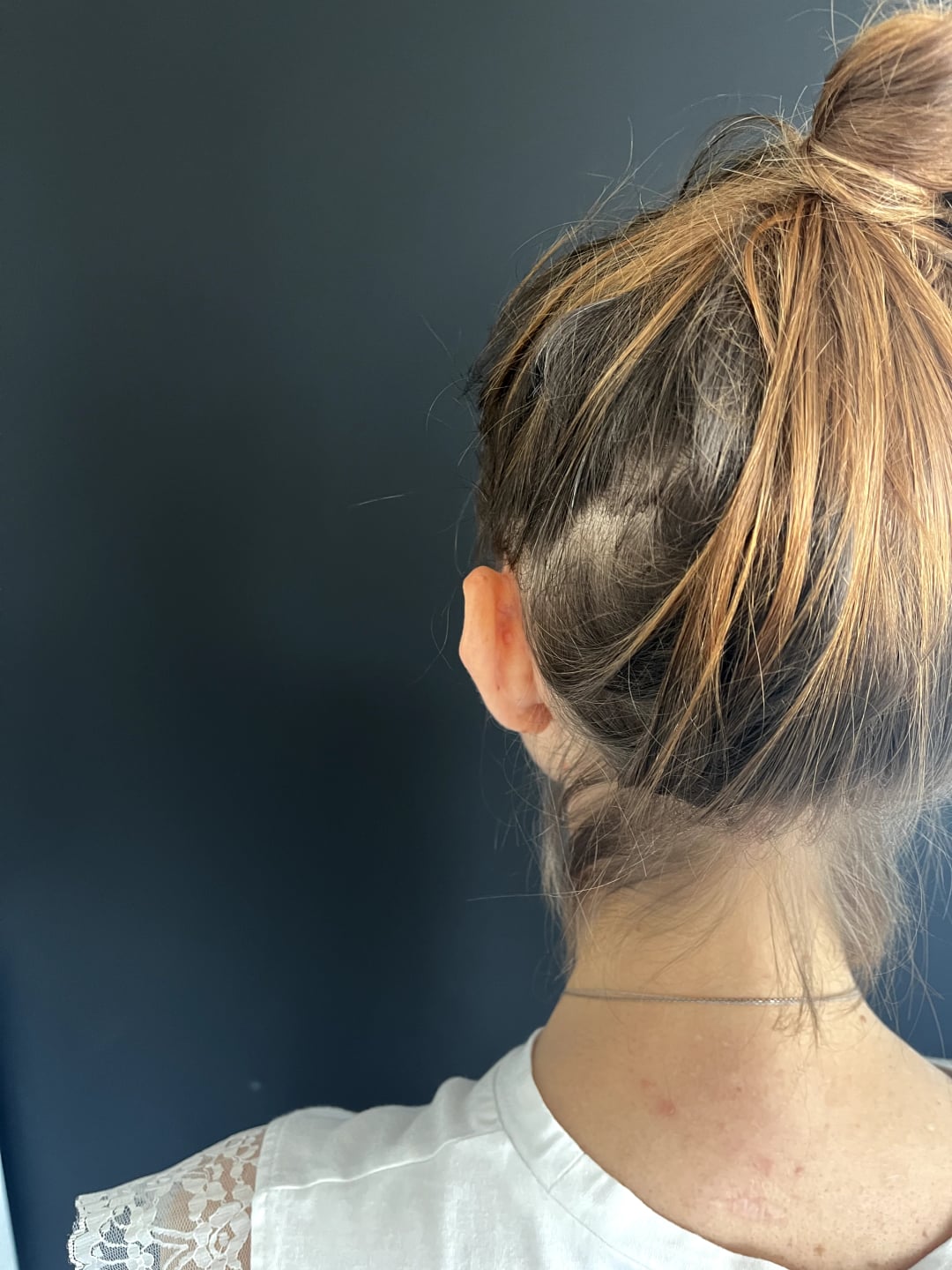 A woman with blonde highlights in her hair stands in front of a dark blue wall, a silver chain around her neck.
